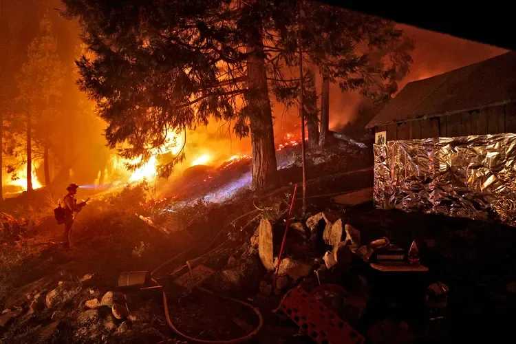 Firefighter defending a foil-wrapped cabin as wildfire burns through the surrounding forest at night.